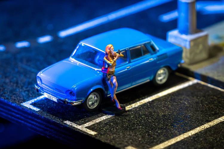 Transport: Cargo – Miniaturmodell eines blauen PKW mit dem Kennzeichen "100 L" und einer Figur einer Geige spielenden Person daneben auf schwarzem Untergrund.