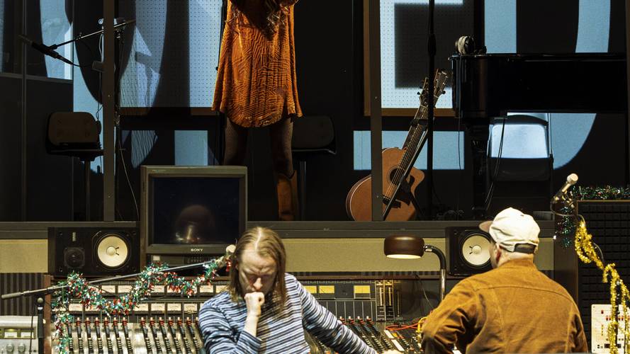 Stereophonic – Person mit langem Haar an Tonmischpult, Weihnachtsdeko und Monitor vorn. Personen in orangefarbenem Gewand im Hintergrund, Gitarren und Klavier. Sony Monitor.