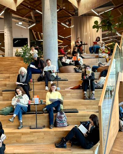 Foto: Pausify – Personen auf Holztribüne in modernem Raum, lesen konzentriert Bücher. Pflanzen und Glaswand im Hintergrund. Entspannte Atmosphäre mit Sitzkissen.