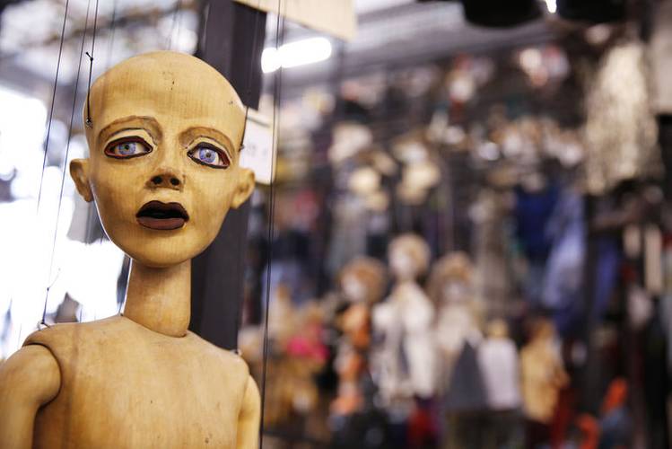 Holzmarionette mit ausdrucksvollem Gesicht, groß im Vordergrund. Hintergrund unscharf mit vielen hängenden Puppen.