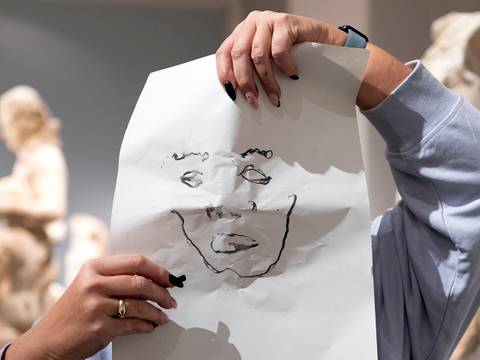 Workshop im Alten Museum – Person hält Blatt Papier mit Gesichtszeichnung; verschwommene Skulpturen im Hintergrund; Fotografie in Innenraum.