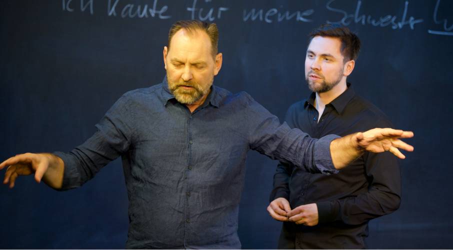 Zwei männlich gelesene Personen vor einer Tafel. Eine Person gestikuliert. Auf der Tafel steht handschriftlich: "Ich kaufte für meine Schwester..."