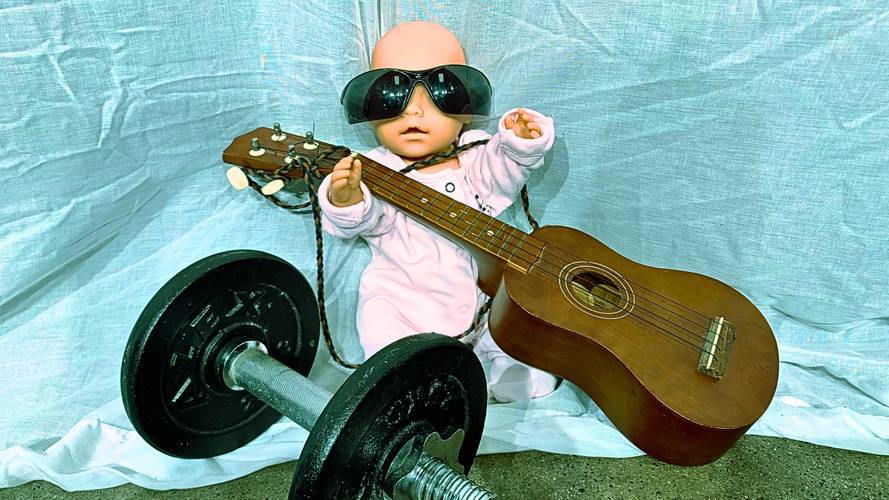 Puppe mit Sonnenbrille haelt Gitarre, sitzt neben Hantel auf Stoffhintergrund.