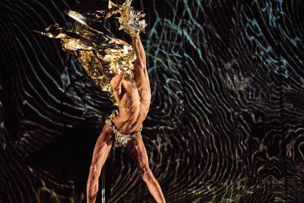 Queer Performance Festival – Person wirbelt mit goldglänzendem Stoff vor dunklem, wellenförmigem Hintergrund.
