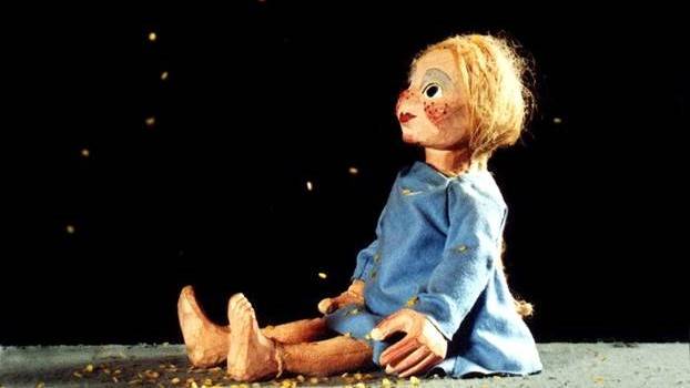Sterntaler – Holzmarionette mit blauem Kleid und geflochtenem Haar sitzt auf grauem Boden; goldenes Konfetti in der Luft schwebend.