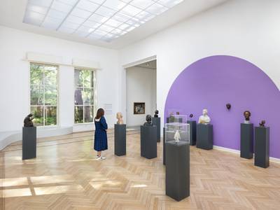 Ausstellungsansicht Georg Kolbe Museum – In einem großen Ausstellungsraum stehen viele dunkle Sockel mit Büsten und Masken mit Abbildern der Schauspielerin Tilla Durieux verteilt. Eine Person schaut sie sich an. Die Wand ist in einem knalligen lila gestrichen.