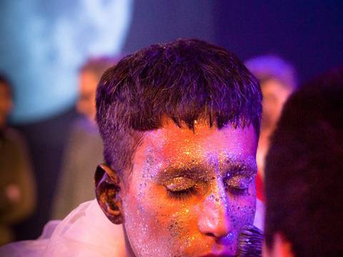  – Person mit Glitzer im Gesicht und geschlossenen Augen trägt weißes, durchscheinendes Gewand. Eine andere Person trägt Goldohrring und pinselt das Gesicht.