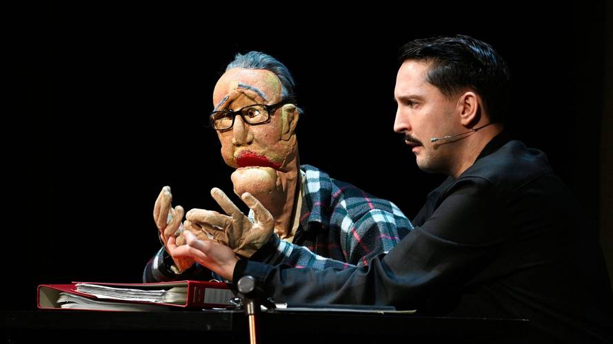 Männlich gelesene Person in Schwarz mit Mikrofon dirigiert Puppe mit Brille und Hemd. Beide hinter rotem Ordner, dunkler Hintergrund.