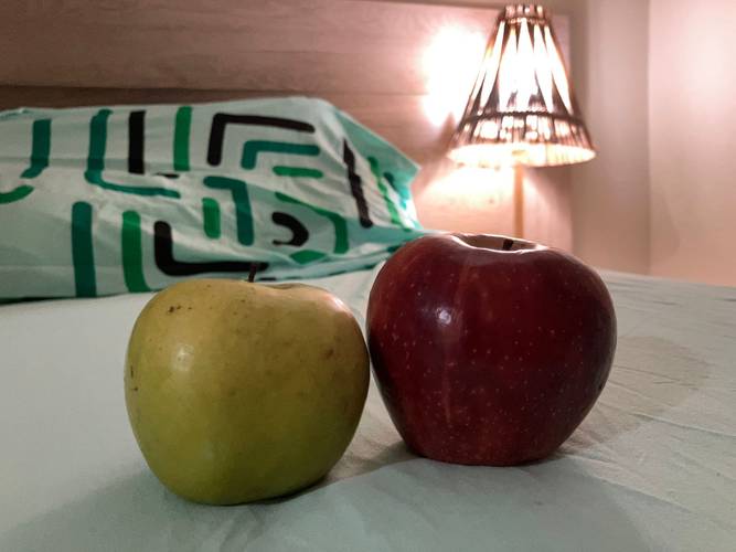 Zwei Äpfel auf einem Bett; ein gelber und ein roter Apfel. Im Hintergrund ein Kissen mit grün-schwarzen Mustern und eine eingeschaltete Lampe.