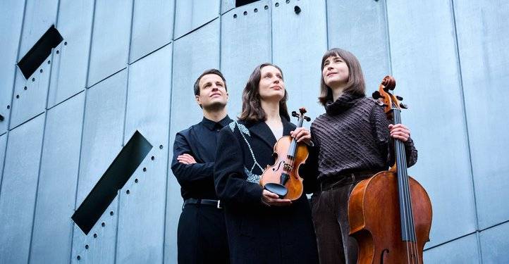 Trio E.T.A. – Drei Personen vor einer Metallwand, mittlere und rechte Person halten Streichinstrumente: Geige und Cello. Alle blicken in die Ferne.