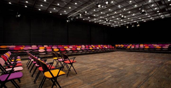 Großer Raum mit Holzboden, umgeben von leeren Stuhlreihen in Pink, Rot und Orange. Decke mit vielen Leuchten und Scheinwerfern.