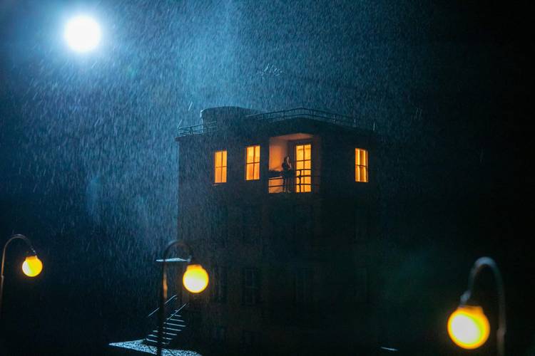 Transport: Departure – Ein Gebäude bei Nacht im Regen, erleuchteter Balkon mit einer Person. Mond am Himmel, vier Straßenlaternen leuchten.