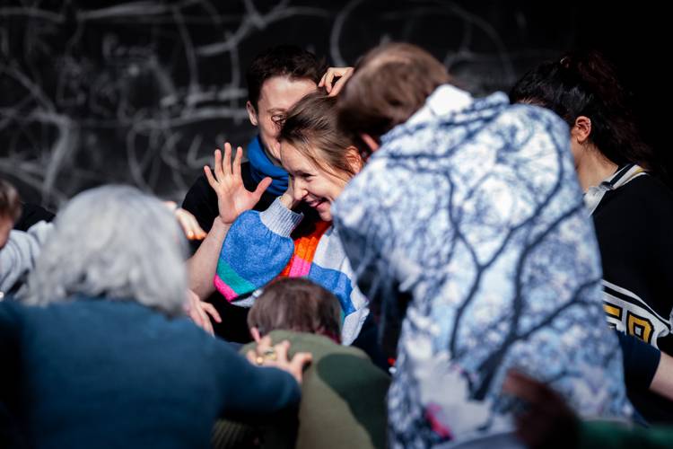 Einblicke: Workshop – Mehrere Personen in lebhafter Unterhaltung; eine Person lacht, trägt einen bunten Pullover. Hintergrund mit dunkler Tafel und weißer Kreidezeichnung.