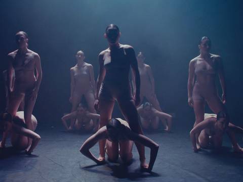 DART Dance Company: KOMPÓT – Personengruppe in Hautfarben-clothing auf Buhne. Zwei stehen, andere knien davor mit Kopf gesenkt. Lichtstrahl von oben beleuchtet Szene dramatisch.