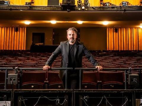 James Gaffigan – Person im schwarzen Anzug lehnt an der Absperrung eines leeren, beleuchteten Theatersaals mit orangefarbenen Vorhängen und leeren Sitzen.