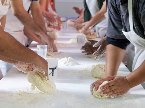 Tiziano Cruz_Bread Workshop – Mehrere Menschen kneten Teig auf einem langen Tisch. Alle tragen Schürzen. Im Hintergrund stehen Becher mit unleserlichem Text.