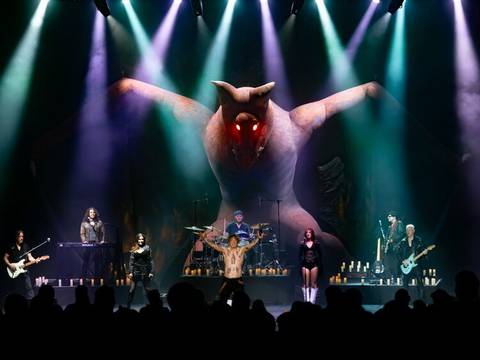  – Bühne mit Band; riesige Fledermausfigur mit leuchtenden roten Augen im Hintergrund. Mehrfarbiges Scheinwerferlicht, viele Kerzen auf der Bühne.