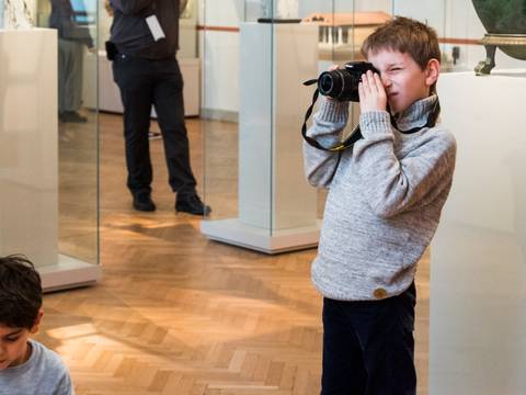  – Kind nimmt in einem Museum mit einer Kamera ein Foto auf. Zweites Kind sitzt am Boden. Hinter ihnen Personen und Ausstellungsstücke in Glasvitrinen.