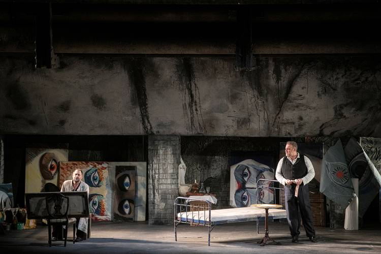 La bohème – Zwei männlich gelesene Personen in einem Raum mit Gemälden von Augen; eine sitzt, die andere steht neben einem Bett.