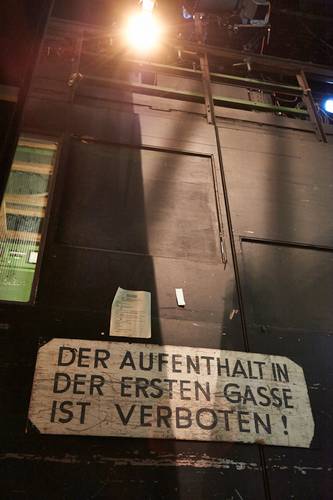 Backstage-Führung – Großes Schild an dunkler Wand mit Text: "DER AUFENTHALT IN DER ERSTEN GASSE IST VERBOTEN!" Oben helle Beleuchtung und einige Metallstreben.
