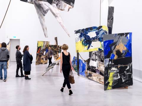 Installationsansicht „Emilio Vedova. Absurdes Berliner Tagebuch ’64“, 1964, Berlinische Galerie © Foto: Lutz Bertram, © Fondazione Emilio e Annabianca Vedova – Foto von Menschen im Ausstellungsraum zwischen Teilen der Installation. Dabei stehen großformatige, abstrakte Installationsobjekte aus bemalten Holzplatten im Ausstellungsraum. Die Platten sind mit kräftigen Pinselstrichen in Schwarz, Weiß, Grau, Gelb und intensivem Blau gestaltet und in verschiedenen Winkeln im Raum positioniert. Der Boden ist glatt und hell, die Wände neutral und ohne zusätzliche Elemente.