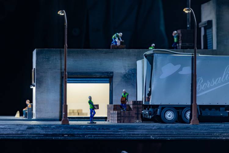 Transport: Cargo – Miniatur-Lagerhaus mit Arbeiter*innen. Personen transportieren Kisten, einige auf einem LKW mit der Aufschrift: "Borsali". Dimmer Innenraum erleuchtet.