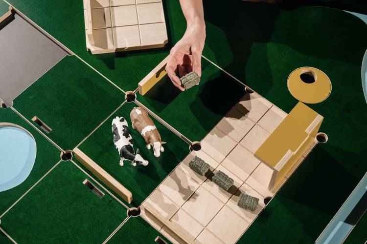 The Game – Miniaturmodell einer Farm mit zwei Kuhfiguren auf grünem Untergrund, Hand hält Miniatur-Heuballen.