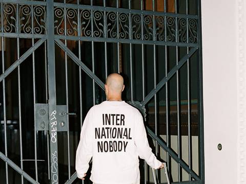 Foto: Lee Everett Thieler – Eine Person mit Glatze, in hellem Outfit, stehen vor einem verschlossenen Tor mit Koffer. Auf dem Ruecken steht "INTERNATIONAL NOBODY".