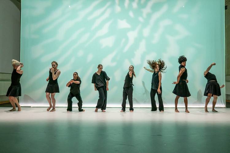 Sieben Personen in schwarzer Kleidung tanzen vor einem hellgrünen Hintergrund. Eine Person wirft ihre Haare in die Luft.