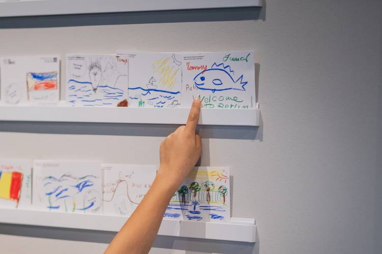 Zeichnungen im Humboldt Labor – Eine Hand zeigt auf bunte Zeichnungen an einer Wand, darunter ein Fisch mit dem Schriftzug "Willkommen in Berlin!".