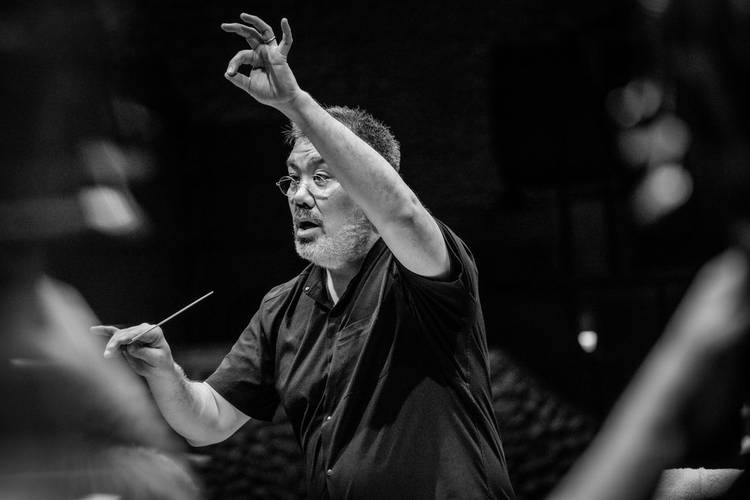 Alan Gilbert – Männlich gelesene Person dirigiert mit einem Taktstock, hebt die Hand, trägt Brille und schwarzes Hemd, konzentrierter Gesichtsausdruck. Schwarzweißbild.