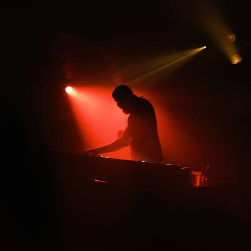 Person an DJ-Pult, umgeben von rotem Licht, mit ausgestrecktem Arm nach rechts. Links oben rote Lampe, rechts zwei weitere Scheinwerfer.