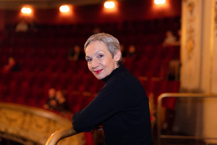 Marion Brasch – Ältere Person mit grauem Haar in schwarzem Oberteil lehnt auf Balkongeländer in leerem Theater mit roten Sitzen und goldener Dekoration.