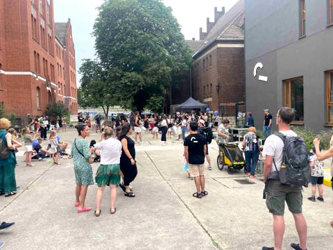  – Menschen auf einem Hof mit roten Backsteingebäuden und Bäumen, ein Kind im Kinderwagen, ein Pavillon im Hintergrund, viele tragen helle Sommerkleidung.