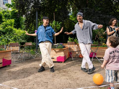  – Jhow und Gissauro führen im Freien eine Hip-Hop-Tanzszene mit offenen Armen auf, während ein Publikum aus Kindern und Erwachsenen aufmerksam zuschaut und mitklatscht.