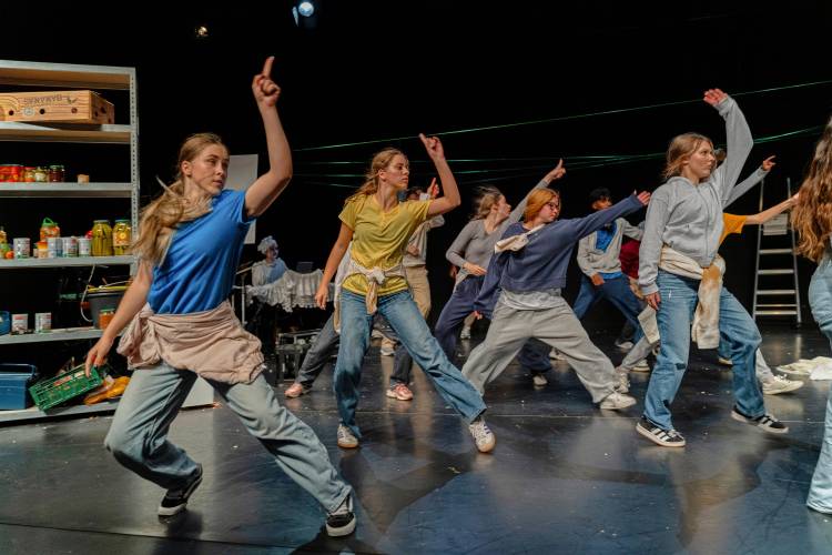 Zum Lachen in den Keller, LABYRINTH Theater Stuttgart – Auf dem Bühnenfoto sind etwa zehn weiblich gelesene Jugendliche abgebildet. Sie tragen Jeans und lockere, bunte Oberteile. Sie wirken, als würden sie gerade tanzen oder sich dynamisch bewegen, da einige Füße leicht in der Luft sind und die Arme ausgestreckt nach oben rechts zeigen. Ihre Körper sind nach vorne gerichtet, sie schauen jedoch ebenfalls nach rechts. Im Hintergrund sind Regale mit Dosen und Kisten sowie eine Leiter zu sehen.