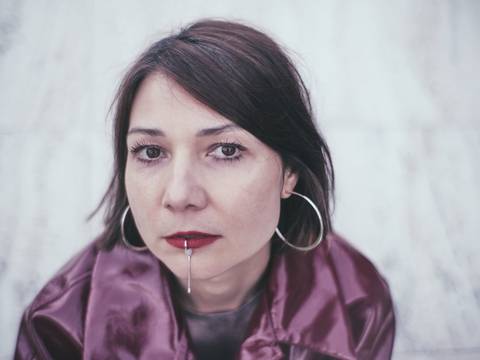 Zuzana Husárová – Person mit Kurzhaarschnitt und großen Ohrringen, trägt glänzenden Mantel, hat roten Lippenstift mit silbernem Stab am Mund. Porträt auf neutralem Hintergrund.