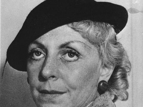 Vicki Baum – Person mit dunklem Barett und hellem, texturiertem Mantel. Kurze, wellige Haare. Die Person blickt ernst. Hintergrund unscharf.