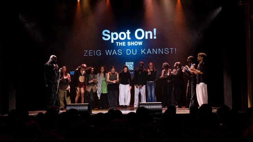 Gruppe von 11 Personen auf Buehne, einige klatschen, andere schauen zur Seite. Hintergrundtext: "Spot On! The Show ZEIG WAS DU KANNST!".