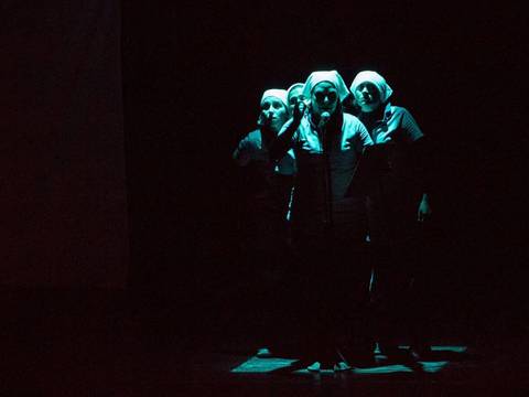  – Fünf Performer*innen stehen dicht beieinander auf einer dunklen Bühne. Sie tragen Arbeitskleidung und helle Kopftücher. Eine Person steht am Mikrofon, die anderen gruppieren sich dahinter. Das Bühnenlicht ist gedämpft und richtet sich auf die Gruppe.