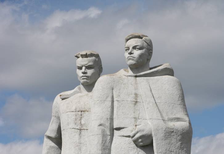 Zwei steinerne Statuen von maennlich-gelesenen Personen, nebeneinander stehend, mit ernstem Ausdruck. Hintergrund aus Wolken und blauem Himmel.