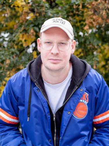Tim Holland – Person mit Brille und Kappe mit Aufschrift "THE NAME IS POETRY", in blauer Jacke mit Basketball-Logo, vor unscharfem Hintergrund mit Bäumen.