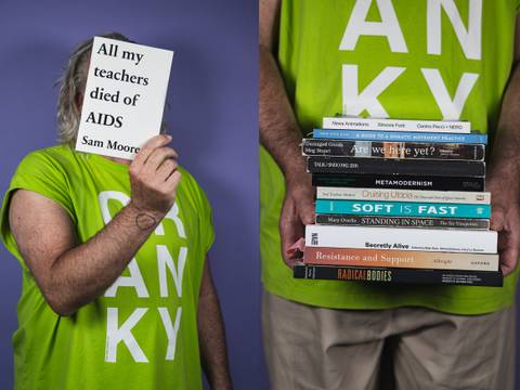 Cranky Choreographing Books – Person mit grauen Haaren in einem grünen T-Shirt mit weißem Schriftzug hält Buch mit Titel "All my teachers died of AIDS Sam Moore" und einen Stapel Bücher.