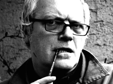 Porträt Wolf Hogekamp, Begründer und Erfinder der Deutschen Poetry-Slam-Meisterschaften – Mann mit grauen Haaren und Brille hält einen Stift im Mund, schaut bedacht und ernst in die Kamera. Schwarz-weiße Fotografie.
