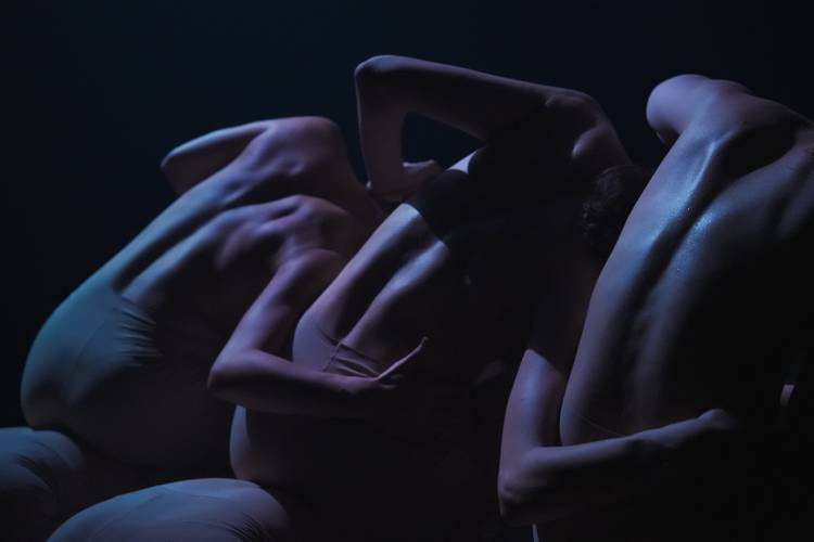 DART Dance Company: KOMPÓT – Drei Personen in dunkler Beleuchtung, Oberkörper unbedeckt, eng zusammengebückt mit ineinander verschränkten Armen und Rücken im Fokus.