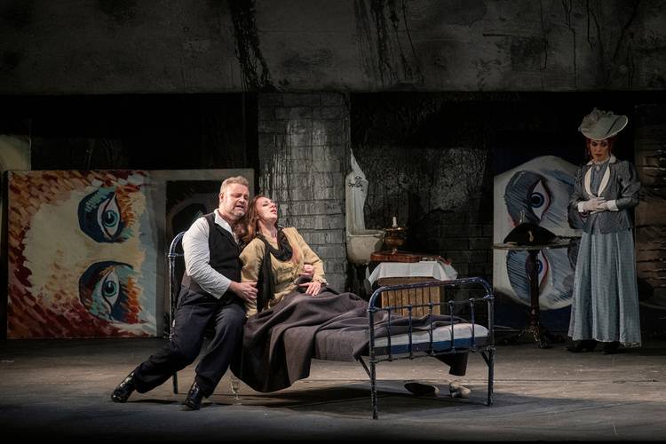 La bohème – Drei Darsteller*innen auf einer Bühne, eine weiblich gelesene Person liegt im Bett, ein männlich gelesener sitzt daneben, im Hintergrund ein Gemälde mit Augen.