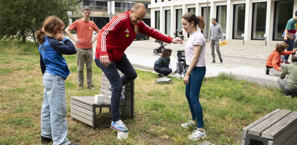 Veranstaltung im Hamburger Bahnhof – Nationalgalerie der Gegenwart – Person in rotem Trainingsanzug balanciert auf Holzbock, zwei Personen beobachten. Weitere Personen im Hintergrund auf Wiese und gepflastertem Weg.