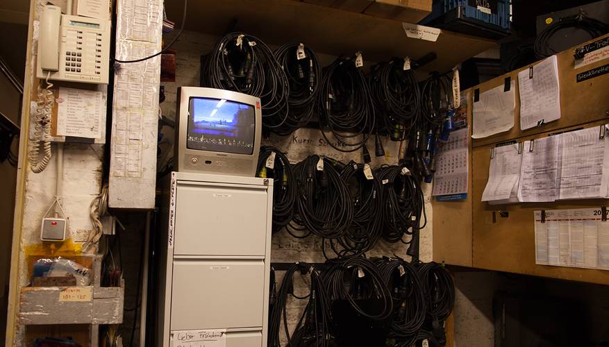 Backstage-Führung – Mehrere Kabel an Wandhaengen, ein Roehrenmonitor zeigt ein Landschaftsbild, links ein Telefon. Zettel und Kalender an Waenden, Schrank darunter.
