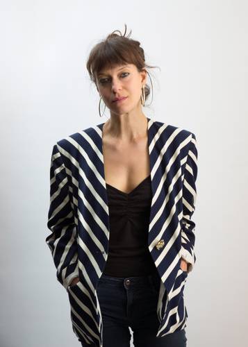 Katharina Bach – Halbporträt einer Person mit langen braunen Haaren im Zopf, trägt einen gestreiften Blazer.