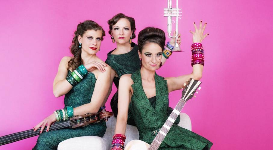 Zucchini Sistaz „Eleganz oder gar nicht!“ – Drei Personen in grüner Kleidung vor pinkem Hintergrund. Eine hält eine Trompete, eine andere eine Gitarre. Alle tragen viele bunte Armreifen.